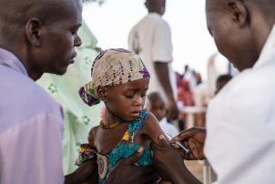 A man supports a small child, while a health care worker gives her a vaccination.