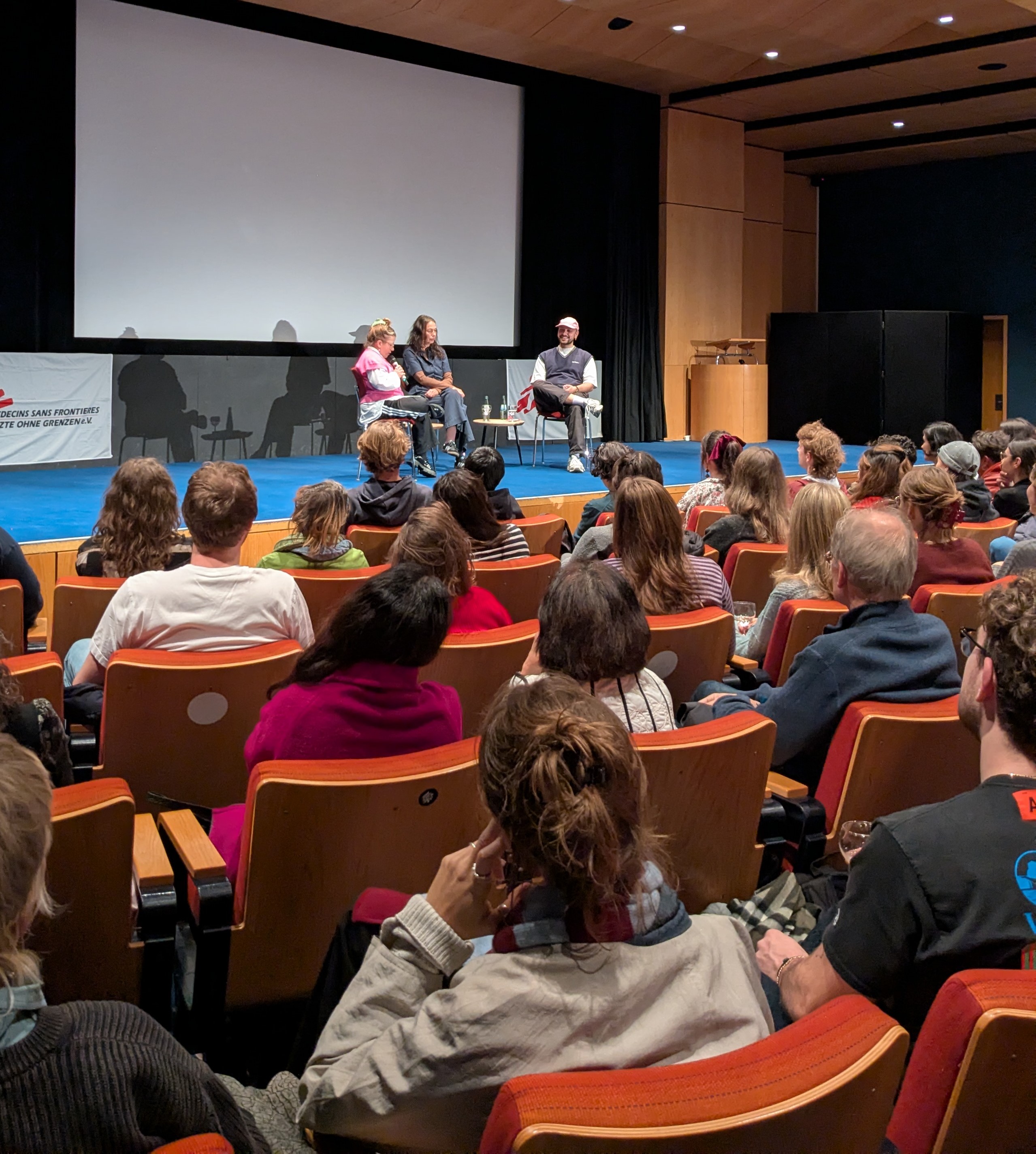 Blick aus dem Publikum auf drei Personen auf der Bühne während eines Live-Gesprächs. Der Saal wirkt voll.