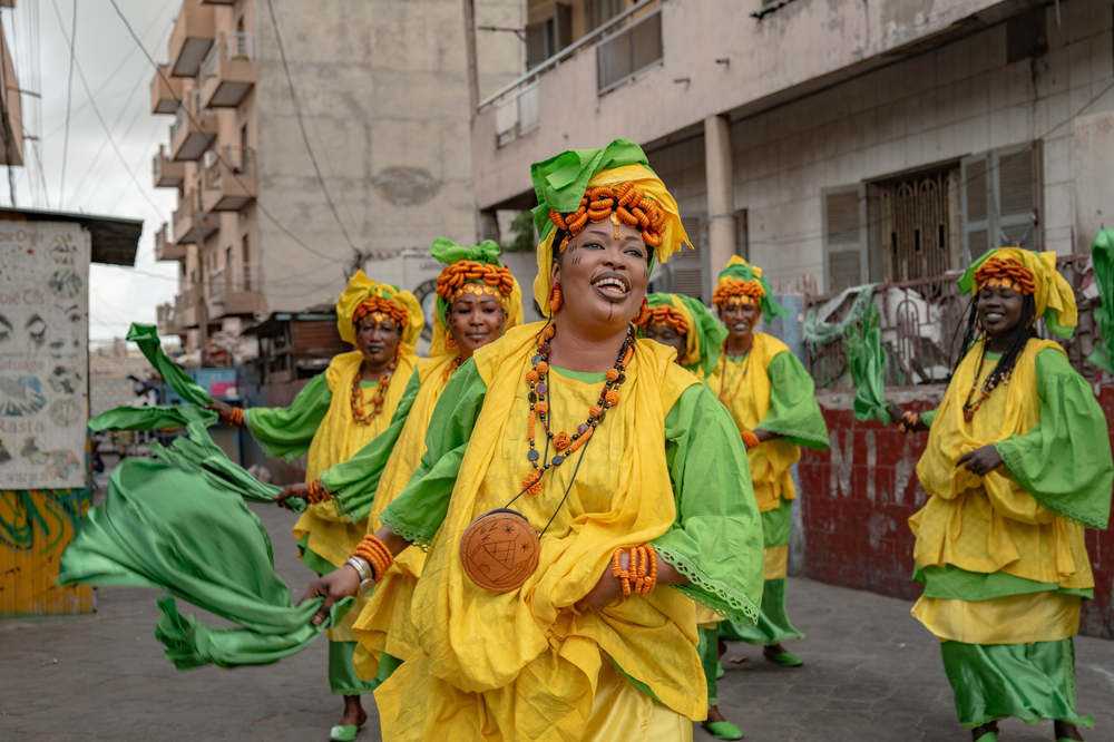 Mehrere Frauen in gelb-grünen Gewändern tanzen und bewegen Tücher auf einer Straße in Dakar.