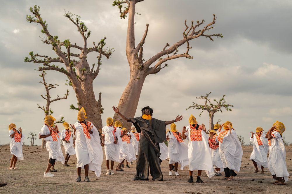 Eine Gruppe von Menschen in weißen Gewändern tanzt vor großen Bäumen, während ein Mann in der Mitte die Arme ausbreitet.
