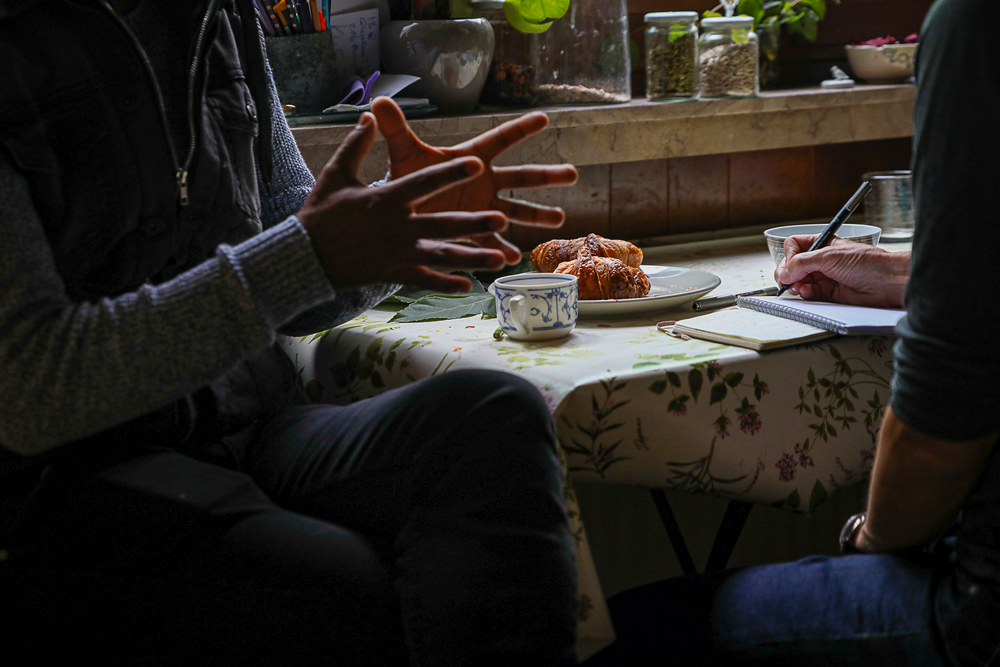 Fotografie zweier Personen, die an einem mit Tassen und Gebäck gedecktem Küchentisch sitzen. Es sind nur die Hände der beiden Personen sichtbar, die links sitzende gestikuliert und die rechts schreibt mit. Interviewsituation