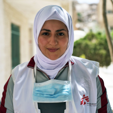 Portraitbild von Ola Barakat. Sie lächelt freundlich in die Kamera und trägt ihre Arbeitskleidung mit Ärzte ohne Grenzen Logo.