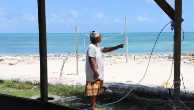 Eine Frau zeigt, wie sich der Strand vor ihrem Haus verschmalert. Am Horizont ist das Meer, das ihrem Haus immer näher rückt.