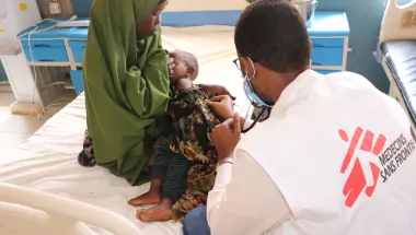 Foto von einer Frau mit ihrem Baby auf dem Arm, welches von einem Arzt untersucht wird. Der Arzt ist von hinten sichtbar und das Ärzte ohne Grenzen-Logo ist auf seinem Gilet aufgedruckt