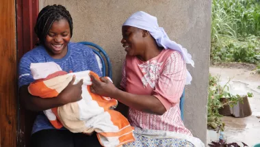 Zwei Frauen sitzen auf einer Bank. Eine hält ein Baby im Arm.