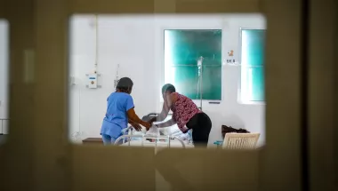 Blick durch verglasten Türrahmen auf Patient*innenbett mit Mitarbeiterin und weiterer Person am Bett