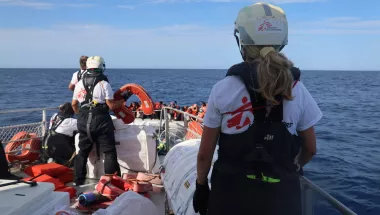 Eine Person steht auf einem Schiff und schaut mit zwei andere Personen geradeaus auf ein kleines Boot mit vielen geflüchteten.