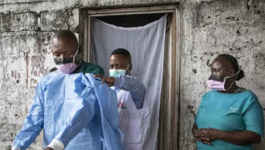 Ein medizinischer Mitarbeiter bekommt einen OP-Kittel angezogen. Eine weitere Mitarbeitern sieht zu