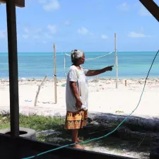 Eine Frau zeigt, wie sich der Strand vor ihrem Haus verschmalert. Am Horizont ist das Meer, das ihrem Haus immer näher rückt.