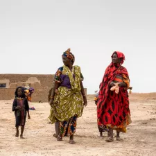 Zwei Frauen in bunten Gewändern, mit jeweils einem kleinen dünnen Kind an ihrer Seite, laufen über trockenen Sand.