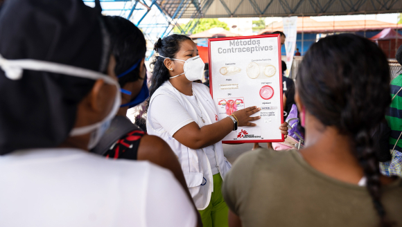 Eine Frau mit Maske spricht vor Anderen, ein Plakat zu Kontrazeptiva in der Hand