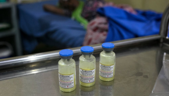 Drei Impfampullen stehen auf einem Tisch in einem Krankenhaus. Im Hintergrund ist eine Patientin zu sehen.