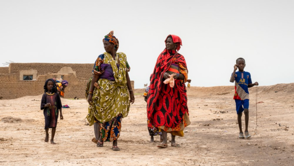 Zwei Frauen in bunten Gewändern, mit jeweils einem kleinen dünnen Kind an ihrer Seite, laufen über trockenen Sand.
