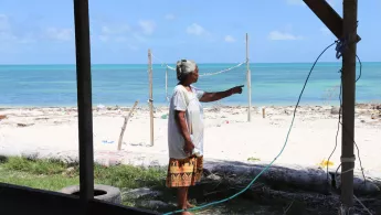 Eine Frau zeigt, wie sich der Strand vor ihrem Haus verschmalert. Am Horizont ist das Meer, das ihrem Haus immer näher rückt.
