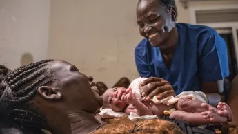 Eine Frau hält ein Baby in den Armen, am Bett steht eine Hebamme