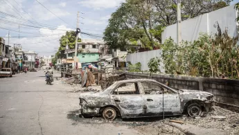 Auf der Straße steht ein ausgebranntes Auto, im Hintergrund fährt jemand auf einem Moped.