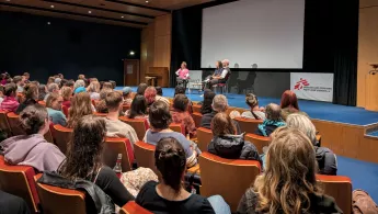 Blick aus dem Publikum auf drei Personen auf der Bühne während eines Live-Gesprächs. Der Saal wirkt voll.