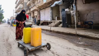 Eine Frau transportiert zwei Wasserkanister auf einem Karren durch eine Straße.