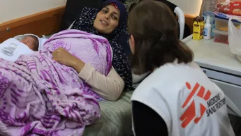 Eine Frau liegt mit Neugeborenem im Bett, eine Mitarbeiterin spricht mit ihr.