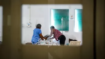 Blick durch verglasten Türrahmen auf Patient*innenbett mit Mitarbeiterin und weiterer Person am Bett