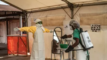 Eine Person in Schutzkleidung wird in einem Zelt von einem Mitarbeitenden mit Desinfektionsspray dekontaminiert.