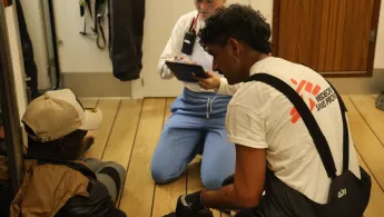 Zwei Mitarbeitende von Ärzte ohne Grenzen sprechen mit einem Mann, der auf dem Boden sitzt und sich an die Wand anlehnt. Ein Mitarbeiter misst den Puls des Mannes.