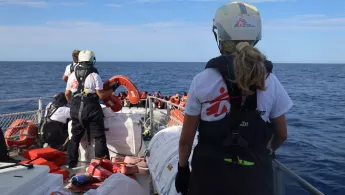 Eine Person steht auf einem Schiff und schaut mit zwei andere Personen geradeaus auf ein kleines Boot mit vielen geflüchteten.