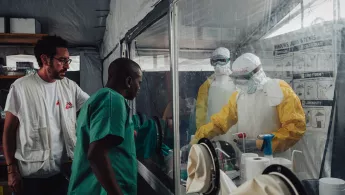 Zwei Personen stehen in Schutzanzügen hinter einer Glasscheibe in einem Labor und interagieren mit zwei medizinischen Mitarbeitern.