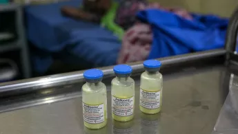 Drei Impfampullen stehen auf einem Tisch in einem Krankenhaus. Im Hintergrund ist eine Patientin zu sehen.