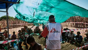 Ein Ärzte ohne Grenzen Mitarbeiter steht vor Patient*innen, die zum Schutz vor der Sonne unter einer aufgespannten Plane sitzen.