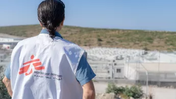 Eine Person von hinten fotografiert, mit einer Ärzte ohne Grenzen Weste die auf ein entferntes Camp schaut