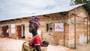 Eine Frau steht mit einem Kind auf dem Rücken vor einem Backsteingebäude mit Logo von Ärzte ohne Grenzen