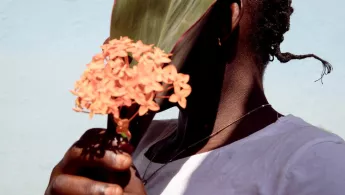 Person die sich ein grünes Pflanzenblatt und eine orangene Blume vor das Gesicht hält um nicht erkannt zu werden 