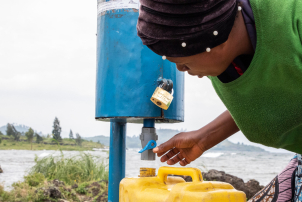 A woman opens a valve in a water system to fill a jug. A wide river flows in the background.