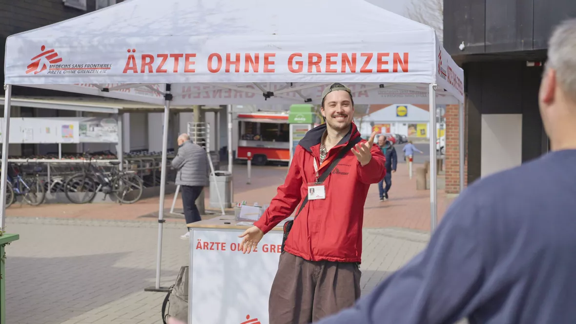 Ärzte ohne Grenzen Mitarbeiter steht unter einem Infostand mit ausgestreckten Arm, während eine andere Person auf ihn zu läuft 