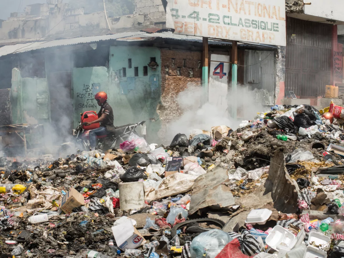 Ein Berg aus Müll und Dreck qualmt auf dem Bild und im Hintergrund ist ein Mensch auf eiem Moped.