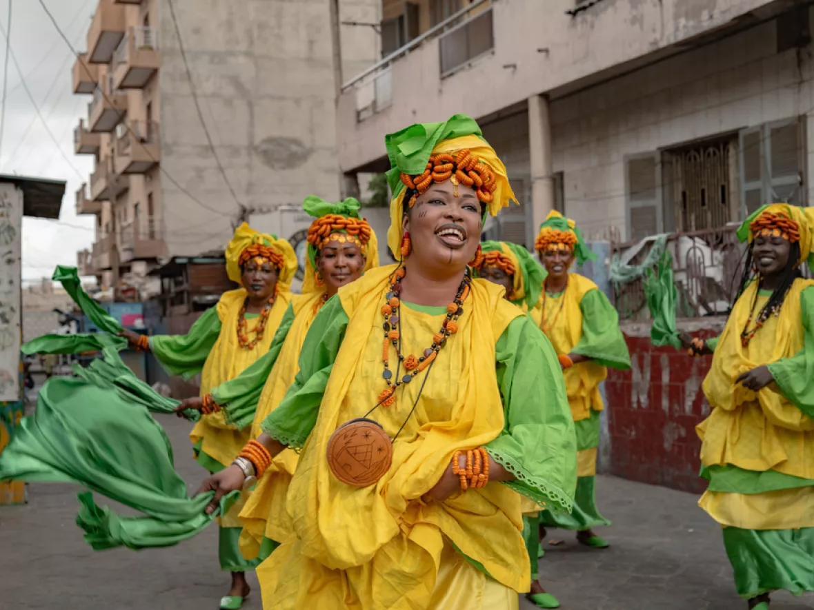Mehrere Frauen in gelb-grünen Gewändern tanzen und bewegen Tücher auf einer Straße in Dakar.