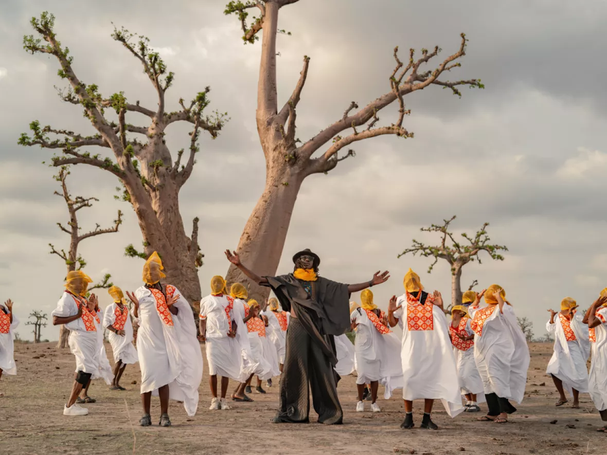 Eine Gruppe von Menschen in weißen Gewändern tanzt vor großen Bäumen, während ein Mann in der Mitte die Arme ausbreitet.