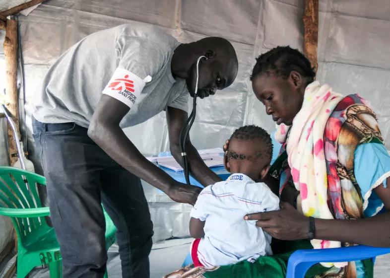Mitarbeiter hört ein Kind mit Stethoskop ab. Daneben die Mutter.