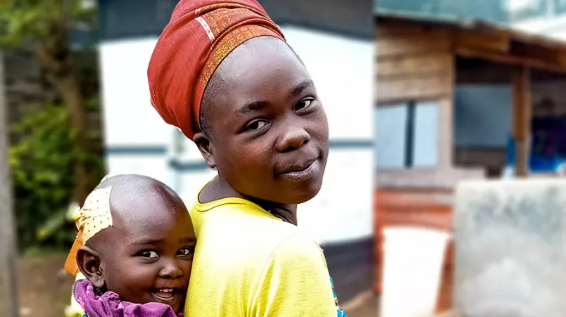 Eine Frau blickt in die Kamera, auf dem Rücken hat sie ein Kleinkind