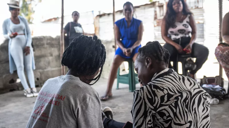 Eine Mitarbeitende spricht mit einer Patien*in. Fünf weitere sitzen gegenüber.