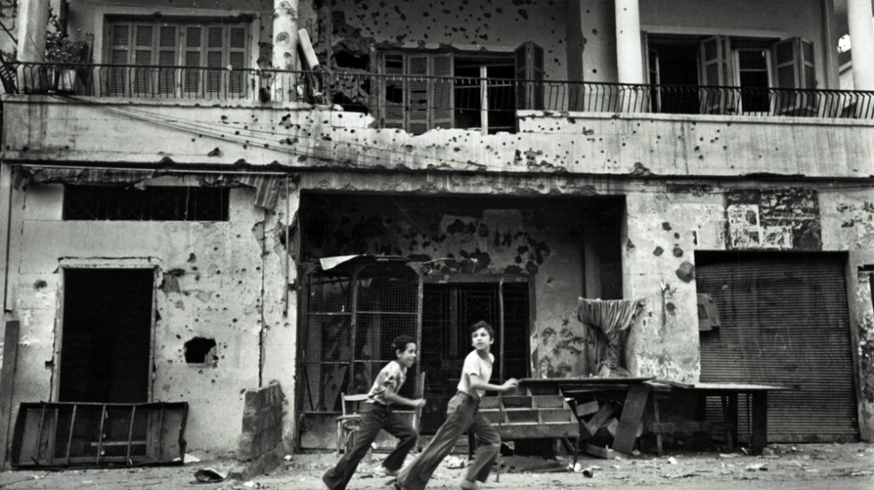 Zwei Jungen rennen durch ein zerstörtes Beirut, hinter ihnen ein Haus mit Einschusslöchern