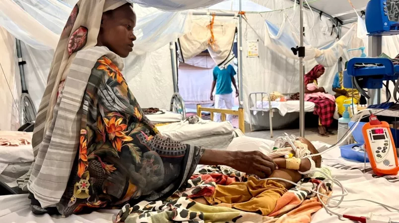 Mutter sitzt mit ihrem Kind auf einem Krankenbett. Das Kind ist an medizinischen Geräten angeschlossen.