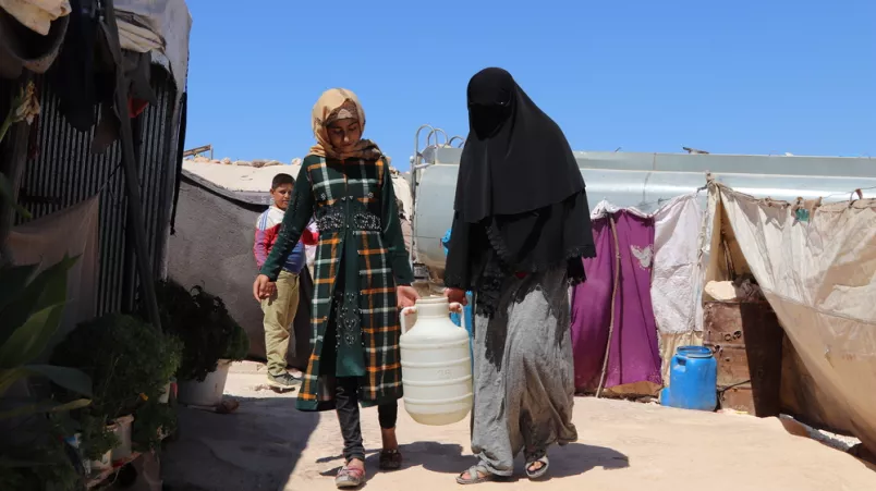 2 Frauen tragen ein großes Gefäß zwischen Zelten.