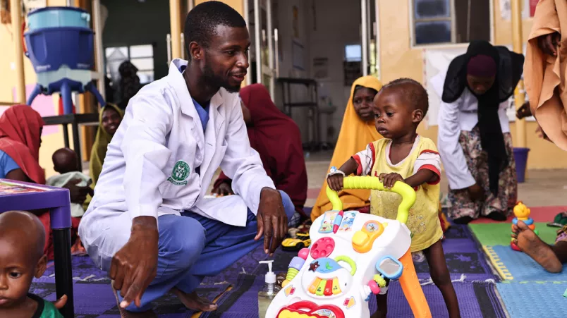 Physiotherapeut hockt neben Kind an Gehhilfespielzeug