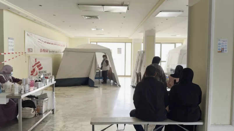 Im Innenraum eines Gebäudes warten Patient*innen auf die Behandlung, daneben eine Ausgabestelle für Medikamente.