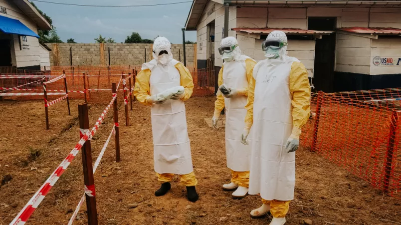 3 Personen stehen in Schutzanzügen vor Absperrbändern im Ebola-Behandlungszentrum