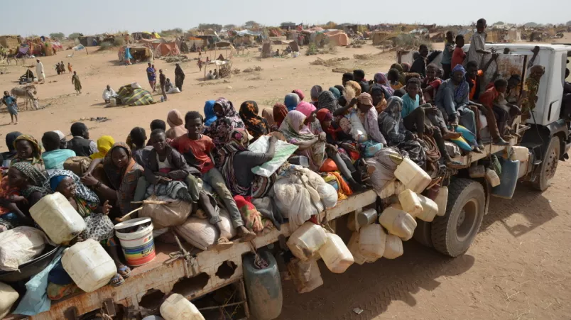 Ein Lastwagen, dessen Ladefläche voller Menschen ist, fährt durch eine Wüstenlandschaft. Die Menschen sitzen zwischen und auf Taschen, Säcken und Kanistern. Im Hintergrund sind Zelte zu sehen.