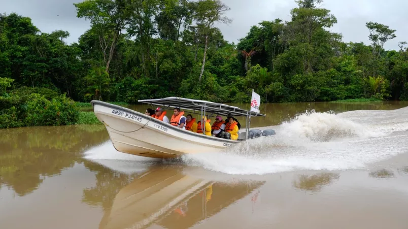 Ein Team fährt mit einem Boot über einen Fluss durch dichtes Waldgebiet. Am Boot ist eine Flagge von Ärzte ohne Grenzen angebracht.