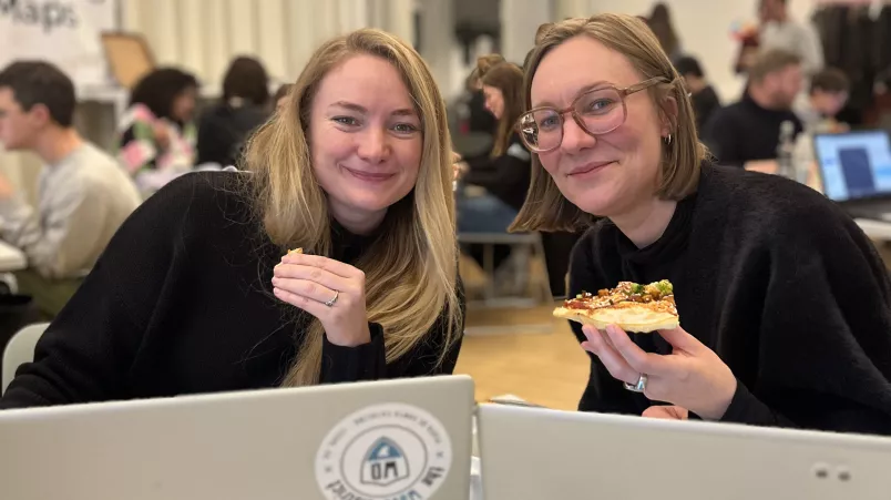 Zwei Personen sitzen vor Laptops und lächeln in die Kamera.